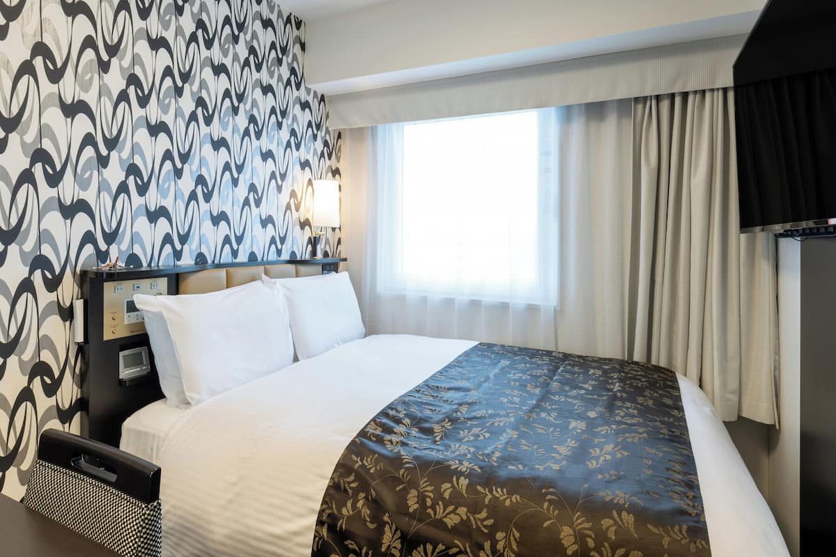 A hotel room with a large bed, a bedside table with a lamp, a TV, and a patterned wall.