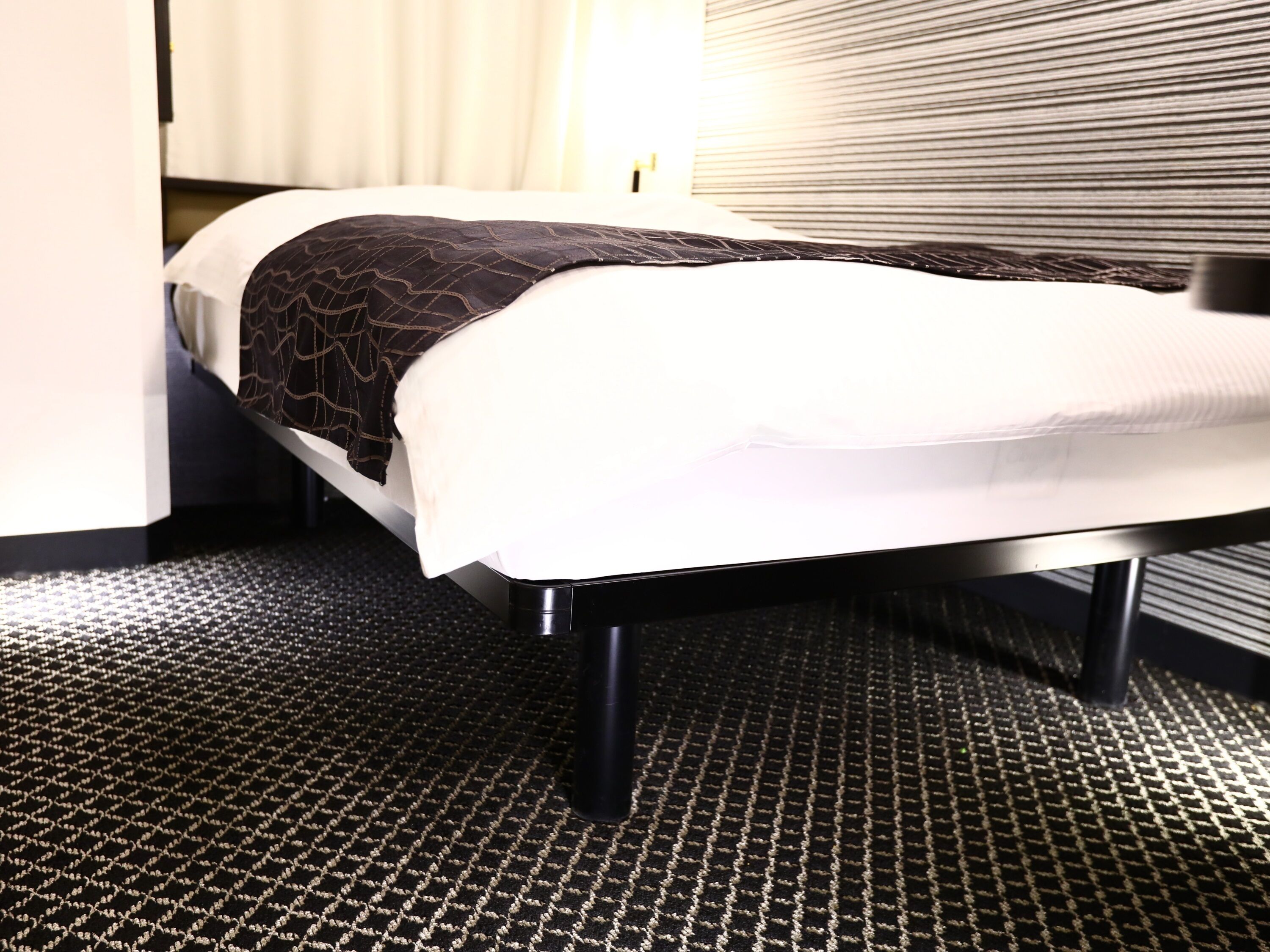 A bed with a white mattress and a dark patterned blanket, a black metal frame, and a black and white checkered carpet.