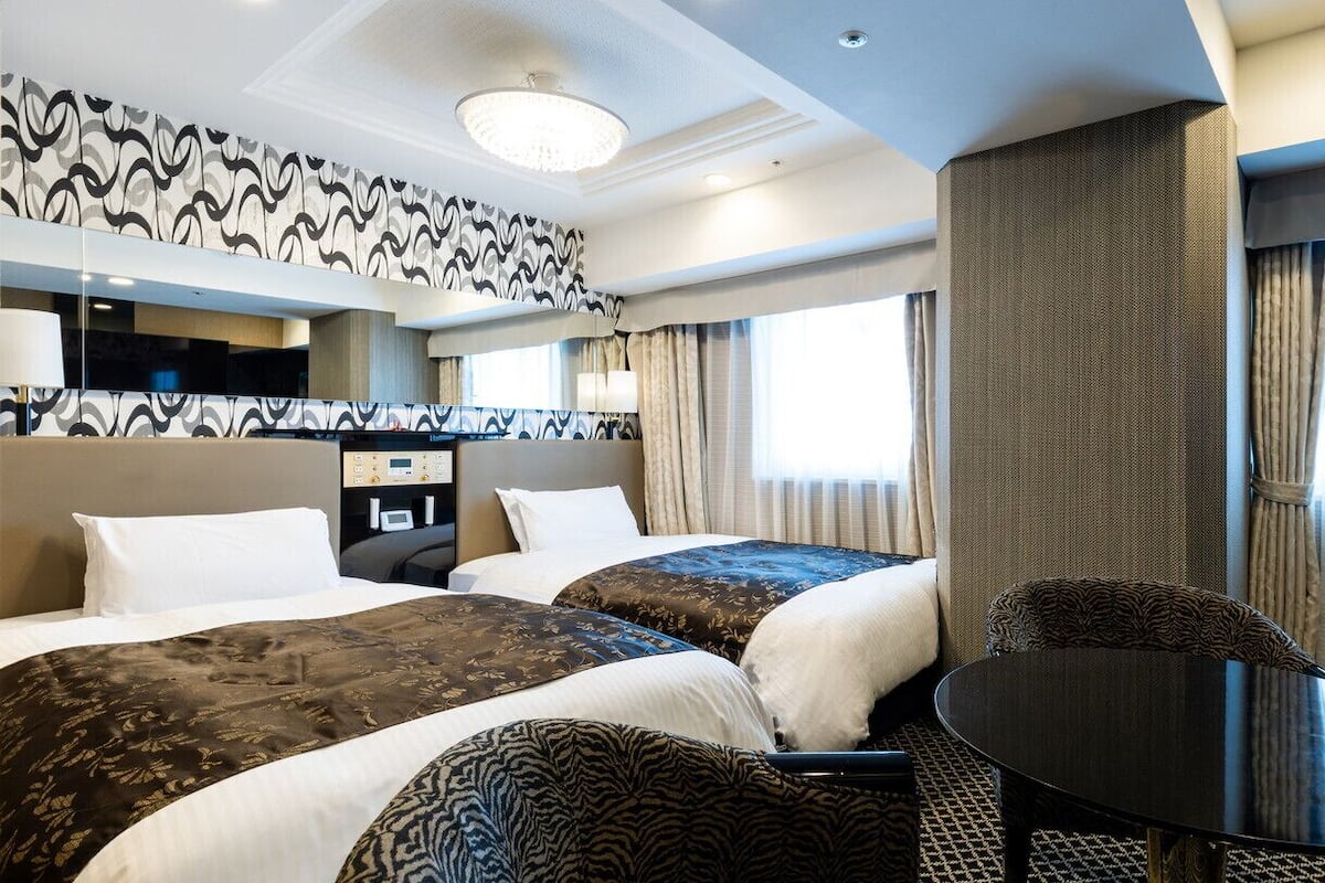 A hotel room with two beds, a small table, a chair, a desk with a computer, and a large window with curtains.