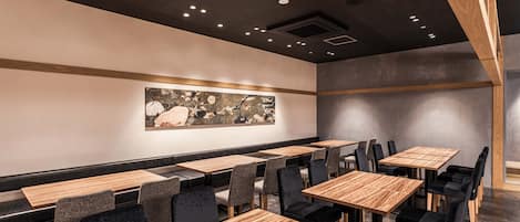 A restaurant interior with wooden tables and chairs, a large abstract painting on the wall, and recessed ceiling lighting.