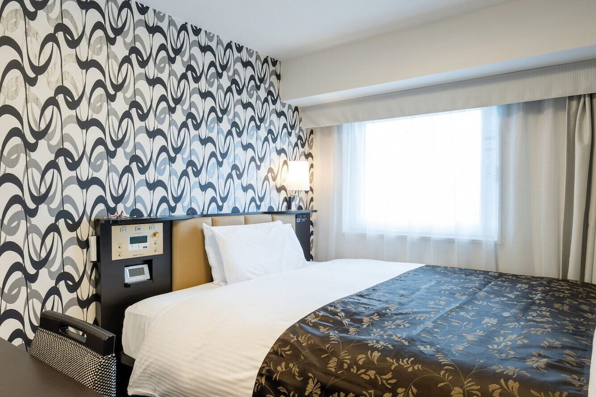 A hotel room with a bed, a control panel, a window with curtains, and a patterned wall.