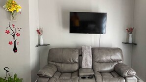 Een woonkamer met een grijze leren bank, een flatscreen televisie aan de muur en twee witte vazen met bloemen op bijzettafels.