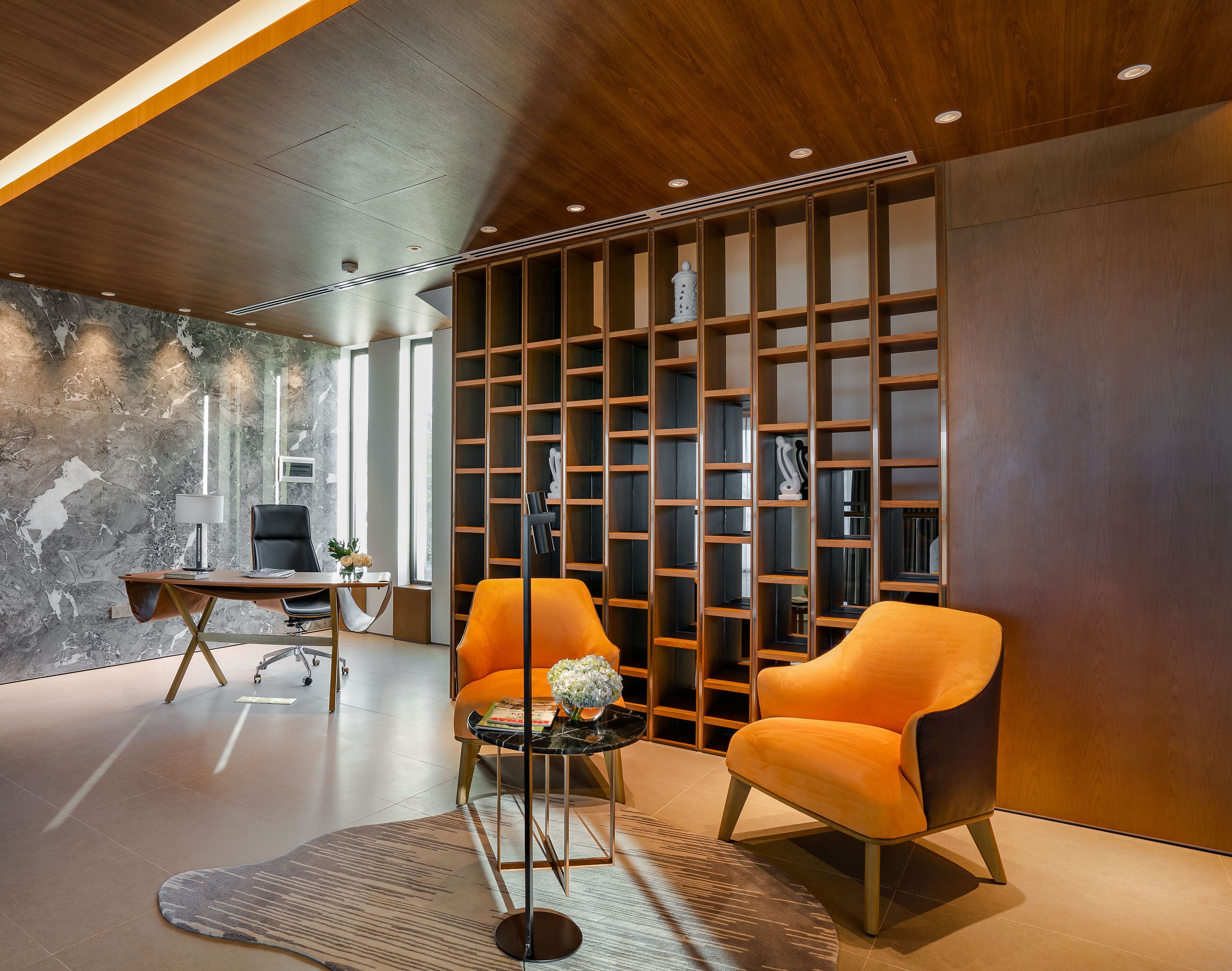 A modern interior with a wooden ceiling, a marble wall, a black leather chair, an orange chair, a small round table with a vase, and a bookshelf.