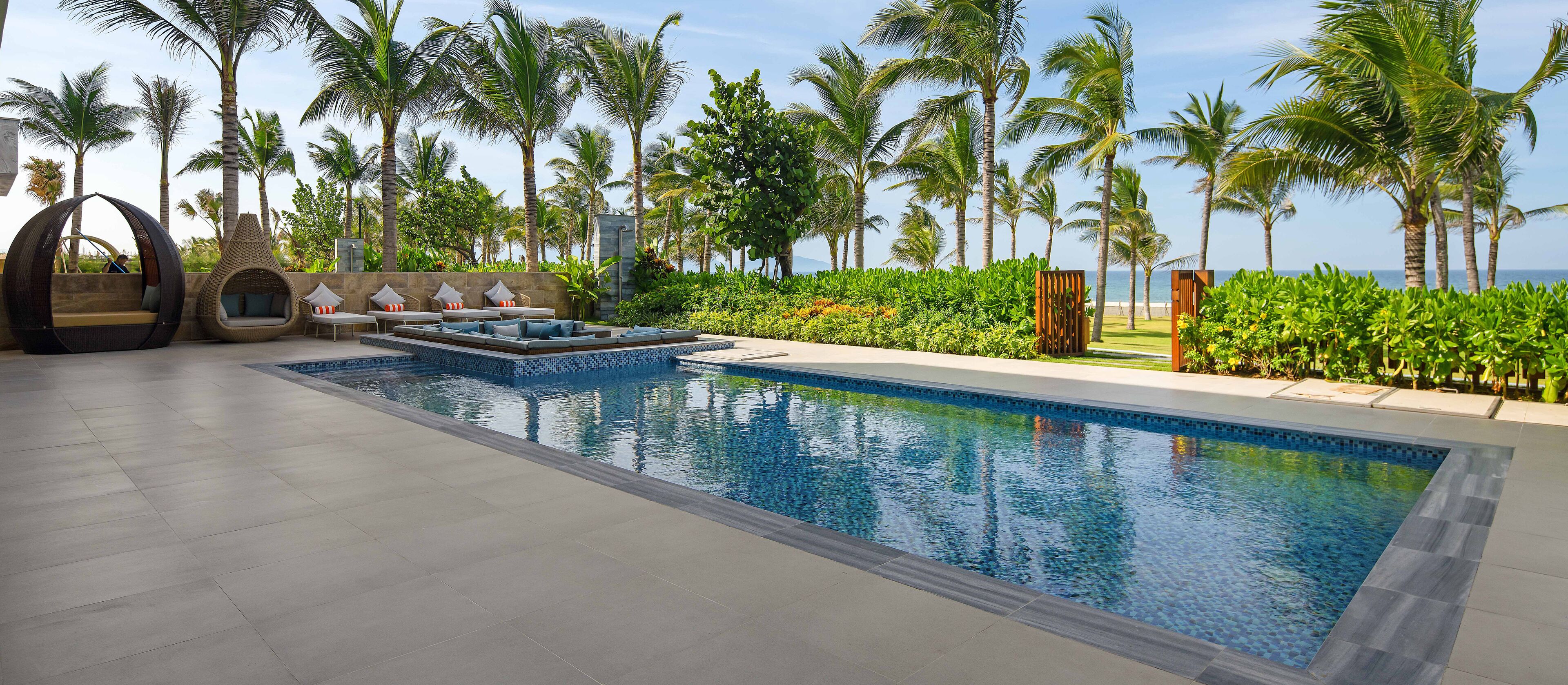 A pool area with lounge chairs and a small seating area, surrounded by palm trees and a view of the ocean.