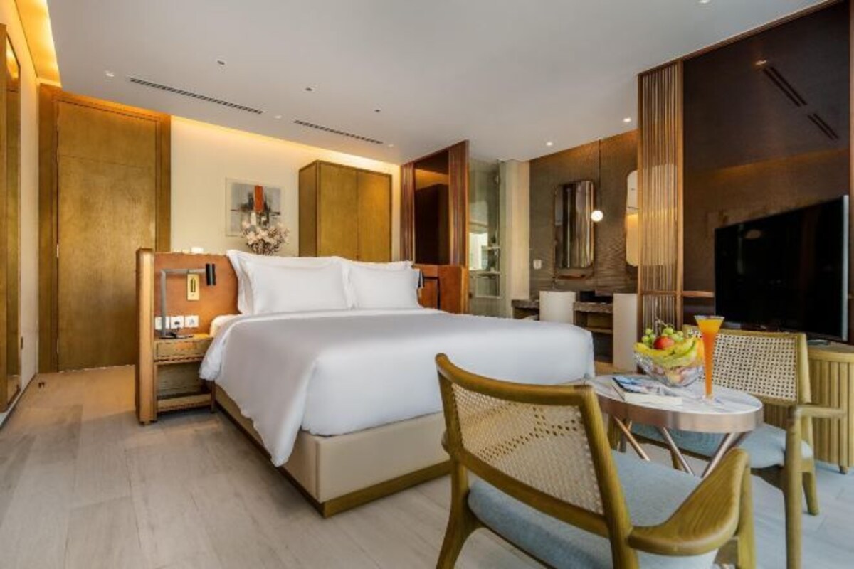 A modern hotel room with a large bed, a bedside table, a chair, a small table with fruit and a drink, and a TV.