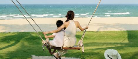 A beach scene with a swing set, a person on a swing, and two bean bags on the grass.