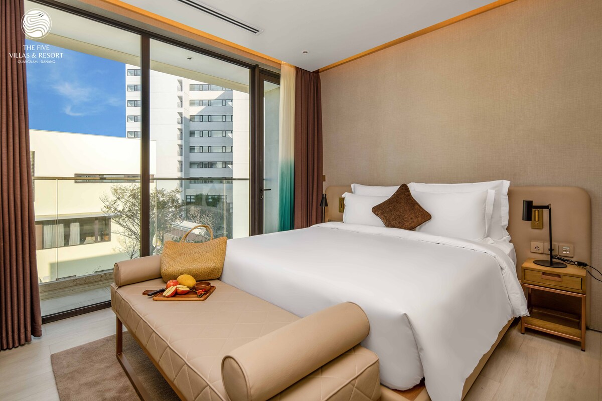 A hotel room with a large bed, a sofa, a nightstand, and a view of the city through a large window.