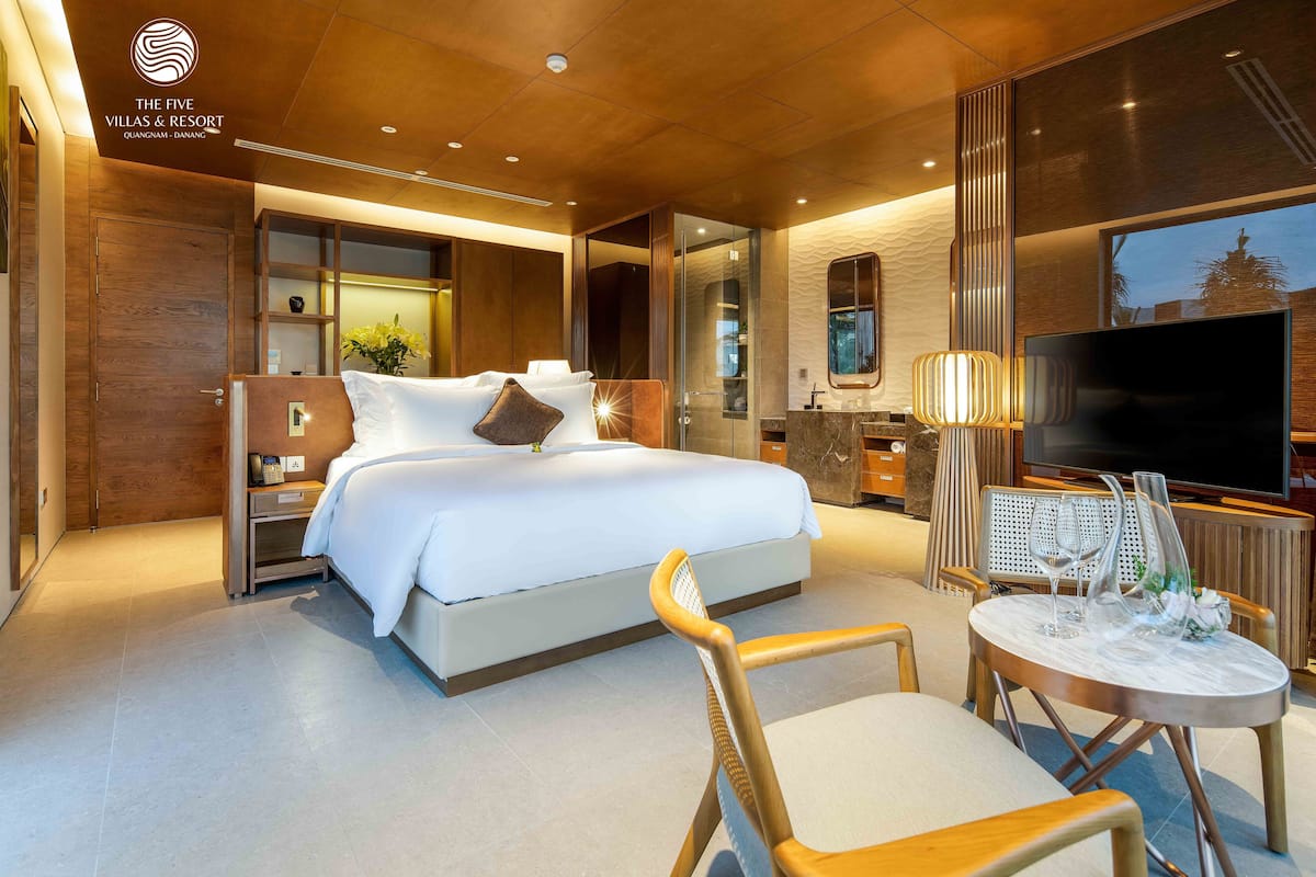 A hotel room with a large bed, a TV, a dining table with chairs, and a bathroom visible through a glass partition.