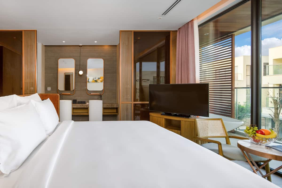 A modern hotel room with a large bed, a flat-screen TV, a dining area with a table and chairs, and a large window offering a view of the city.