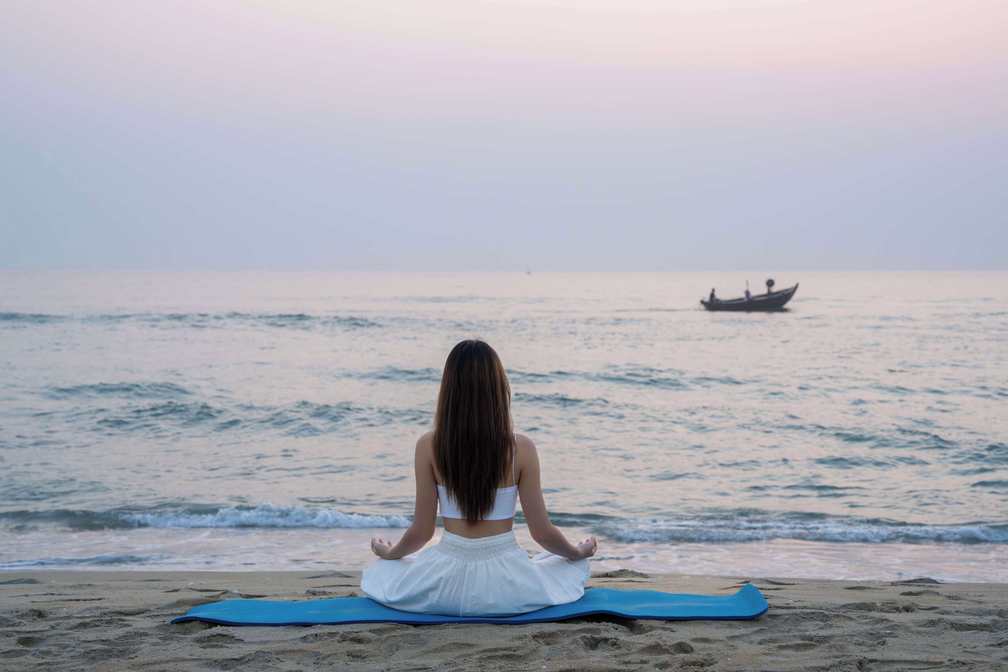 A person sitting on a blue mat on a beach facing the ocean.