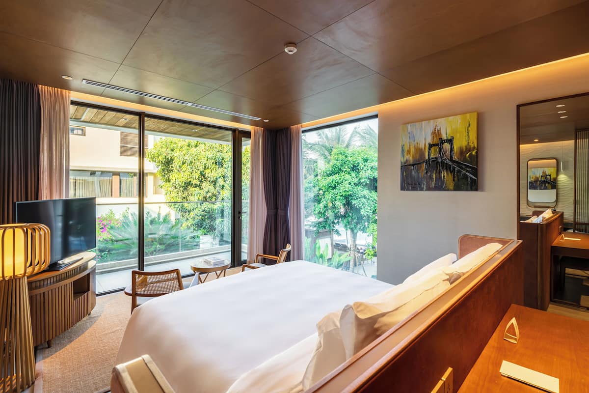 A modern hotel room with a large bed, a TV, a desk, and a window with a view of greenery.