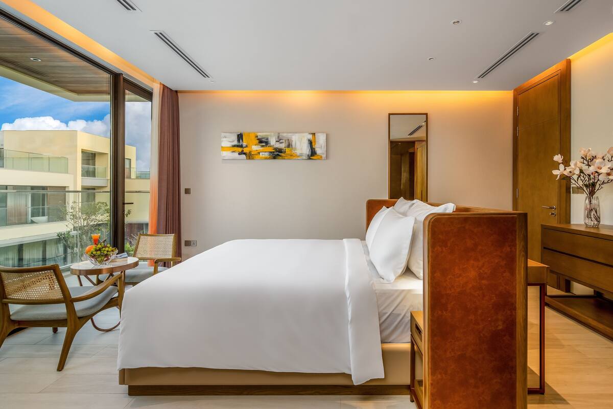 A modern hotel room with a large bed, a desk, a chair, and a small table with fruit. There is a large window with a view of buildings and a painting on the wall.
