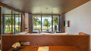 Una habitación de hotel moderna con techo de madera, ventanas amplias con vistas a palmeras y un campo de golf, una mesa de madera con un libro y un florero, y una zona de estar con sofá y sillones.