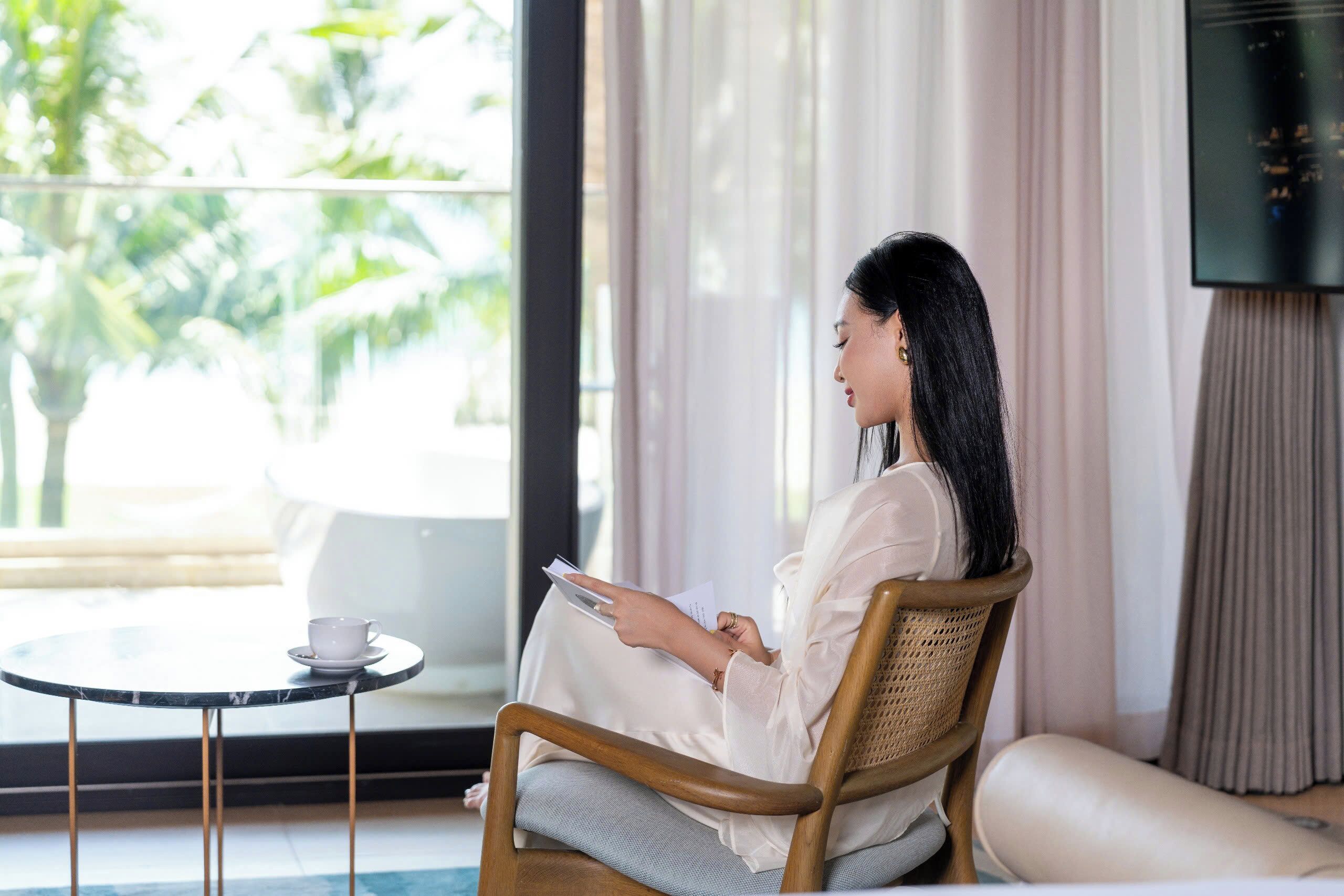 A person sitting on a wooden chair in a room with a view of palm trees through a large window, a small round table with a cup, and a flat-screen TV on the wall.