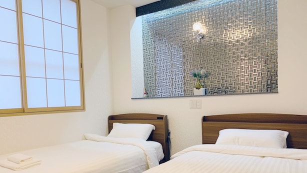 A hotel room with two single beds, a window with a view of a patterned wall, and a small shelf with decorative items. - Allcomein Hotel (Tokyo)