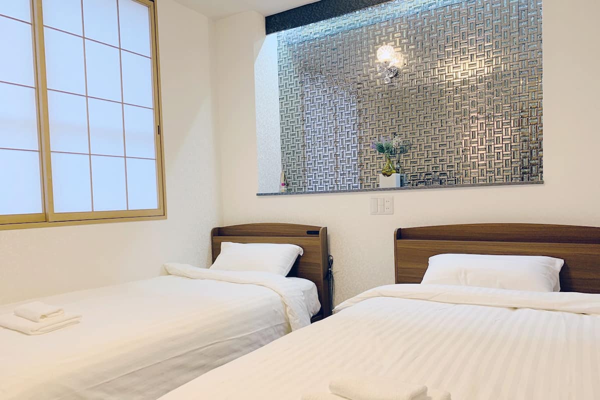 A hotel room with two single beds, a window with a view of a patterned wall, and a small shelf with decorative items.