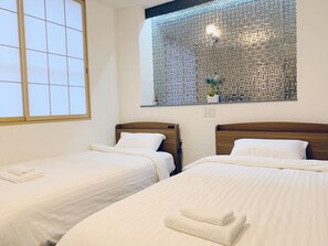 Une chambre d’hôtel avec deux lits simples, une fenêtre donnant sur un mur orné de motifs et une petite étagère avec des objets décoratifs.