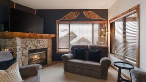 A cozy living room with a stone fireplace, a comfortable sofa, and a television mounted on the wall. - True Ski in/Ski out Hot tub In the village (Big White)