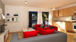 A modern living room with a red sofa, grey couch, and a dining table. There is a kitchen with wooden cabinets and a window overlooking a balcony.
