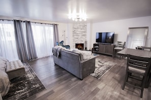 Una sala moderna con sofá, reposapiés y un comedor con mesa y sillas. Un televisor está montado en la pared, y se observa una chimenea.