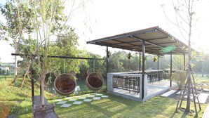 A covered outdoor seating area with hanging chairs and a table, surrounded by greenery and a water feature.