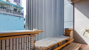 Eine Holzdielenfläche mit Bänken und einem Tisch, eine wellenförmige Metallwand und ein blaues Gebäude im Hintergrund.