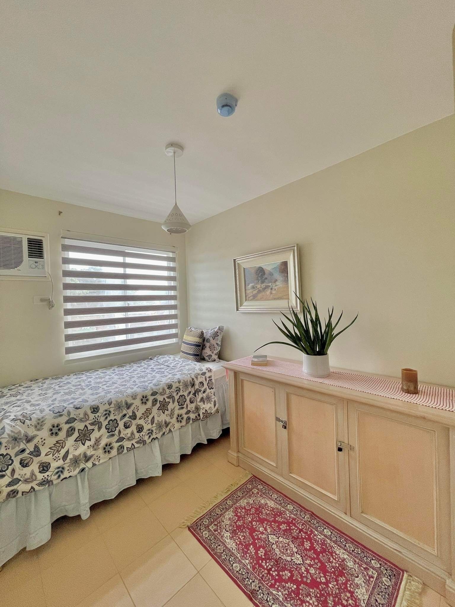 Un dormitorio con cama, mesita de noche, radiador, ventana con persianas, ventilador de techo, un cuadro en la pared y una alfombra roja en el suelo.