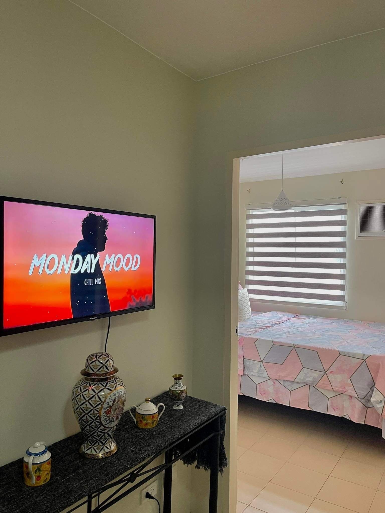 A room with a television displaying 'MONDAY MOOD CHILL MIX', a bed with a geometric-patterned bedspread, a small table with decorative items, and a window with blinds.