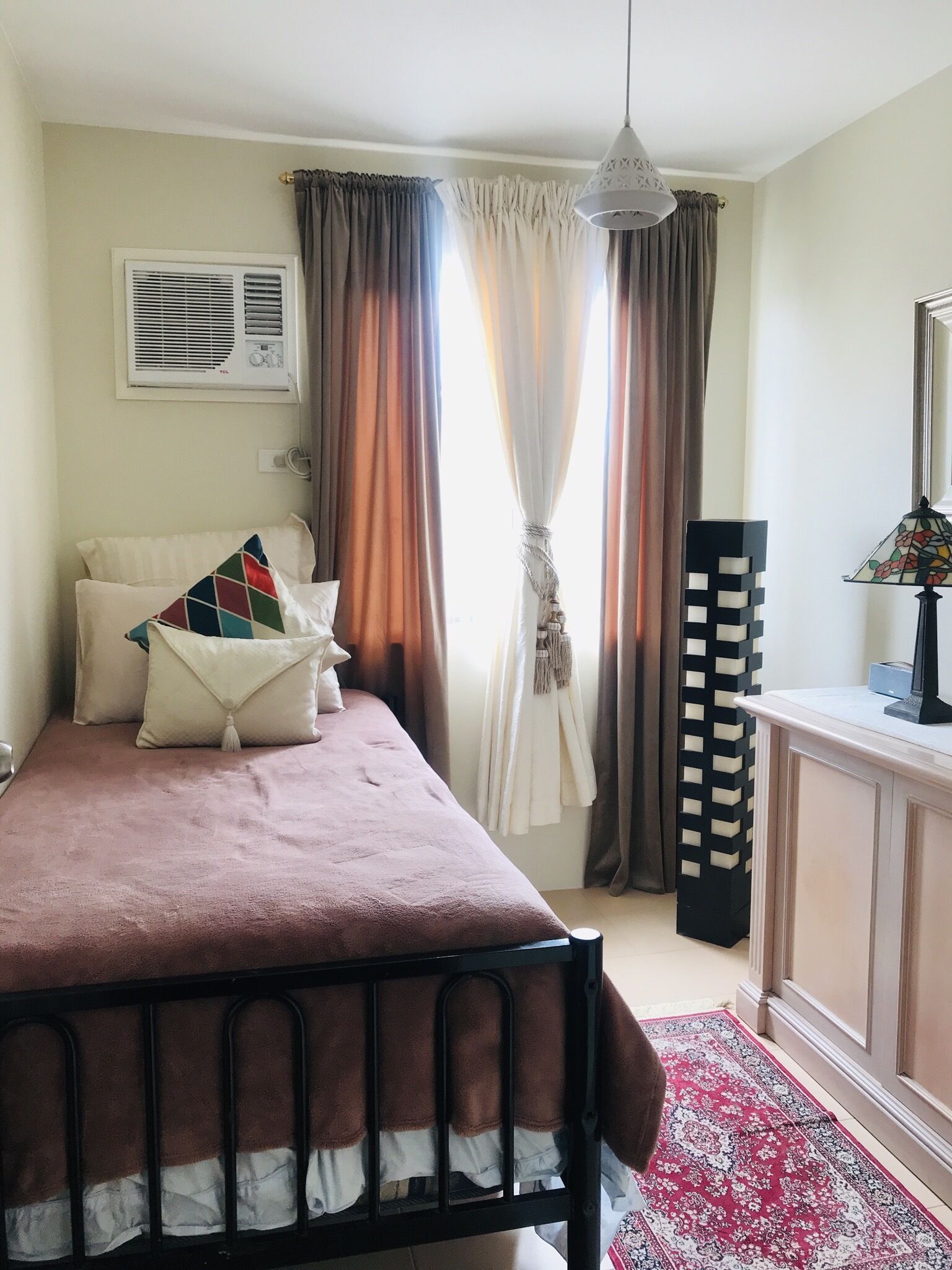 A bedroom with a bed, a radiator, a window with curtains, a hanging lamp, a bedside table, and a patterned rug.