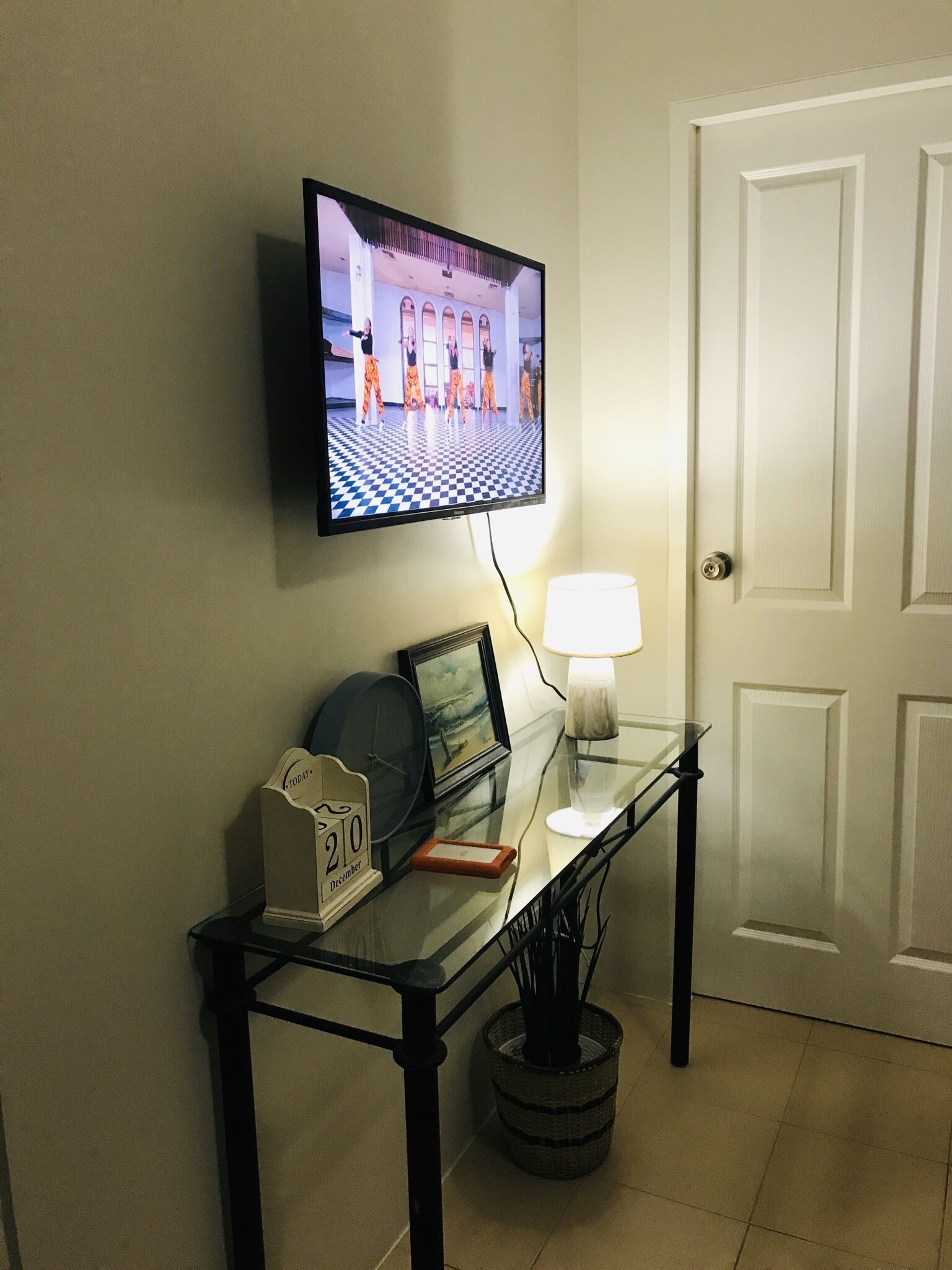 A flat-screen TV mounted on a wall, a glass table with a lamp, a calendar, and a framed picture.
