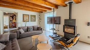 A modern living room with a sofa, two small tables, and a leather chair. A fireplace is built into the wall, and a TV is mounted above it. - Modern villa with heated pool and Jacuzzi, near Ripe, just 20 minutes from the beach of Senigallia (Trecastelli)