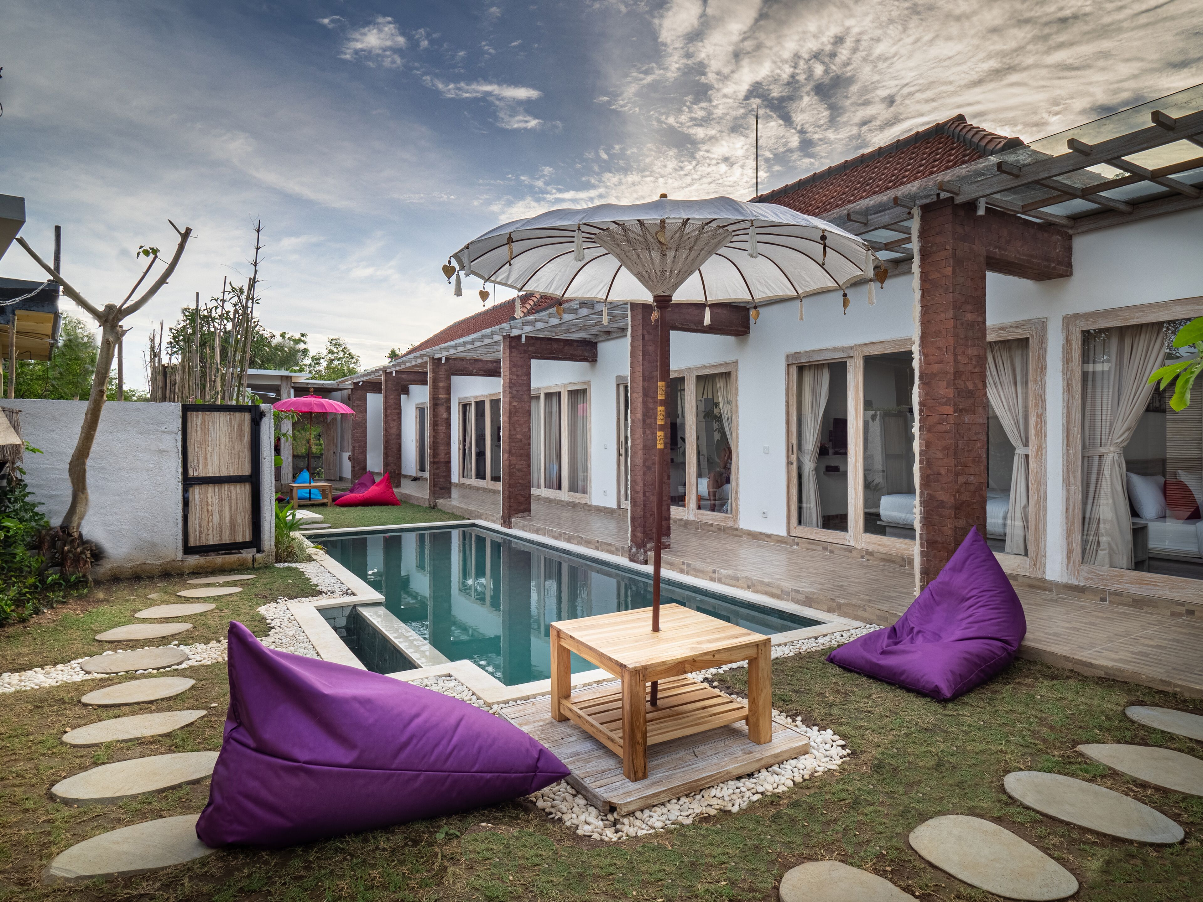 A pool area with a wooden table and purple bean bags, surrounded by a white wall and a pergola with an umbrella.