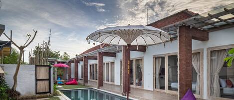 A pool area with a wooden table and purple bean bags, surrounded by a white wall and a pergola with an umbrella.