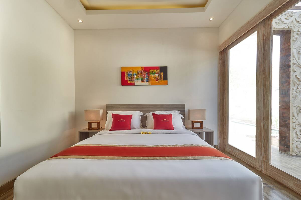 A hotel room with a large bed, two bedside tables with lamps, a colorful painting on the wall, and a large window with a view.