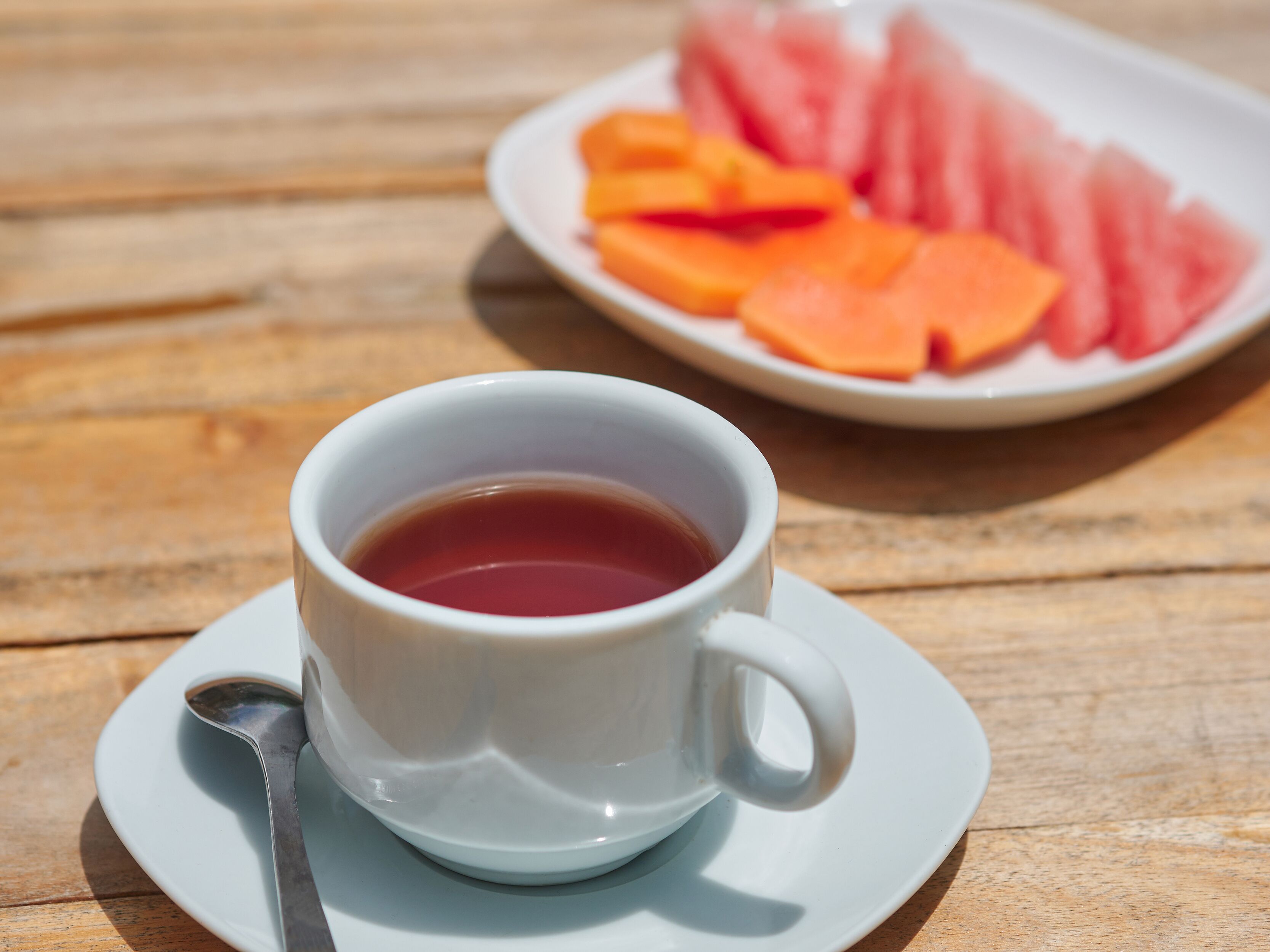A white cup of tea on a saucer with a spoon, and a plate of sliced fruits in the background.