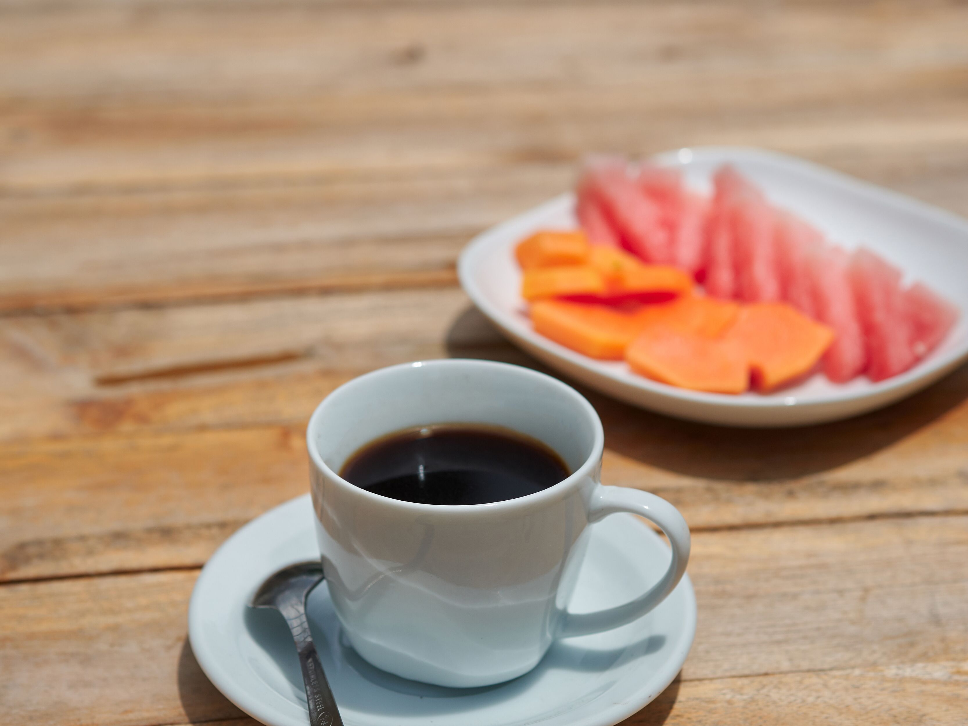 Een witte koffiekop op een schotel met een lepel, en een bord met gesneden fruit op de achtergrond.