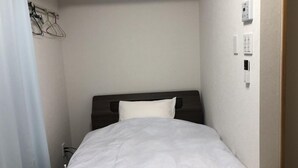 A small, neatly made hotel room with a single bed, a bedside table, and a wall-mounted light switch. - Amami Lucky Life (Amami)