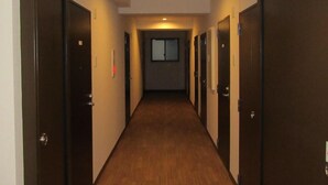 A narrow, well-lit hallway with wooden flooring and dark doors on both sides, leading to a room with a window. - Amami Lucky Life (Amami)