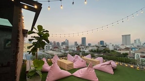 A rooftop terrace with artificial grass, pink bean bags, string lights, and a city skyline view. - The LOCAL Ari - Hostel (Bangkok)