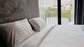 A bedroom with a large bed, a concrete wall, and a view of buildings and greenery outside.