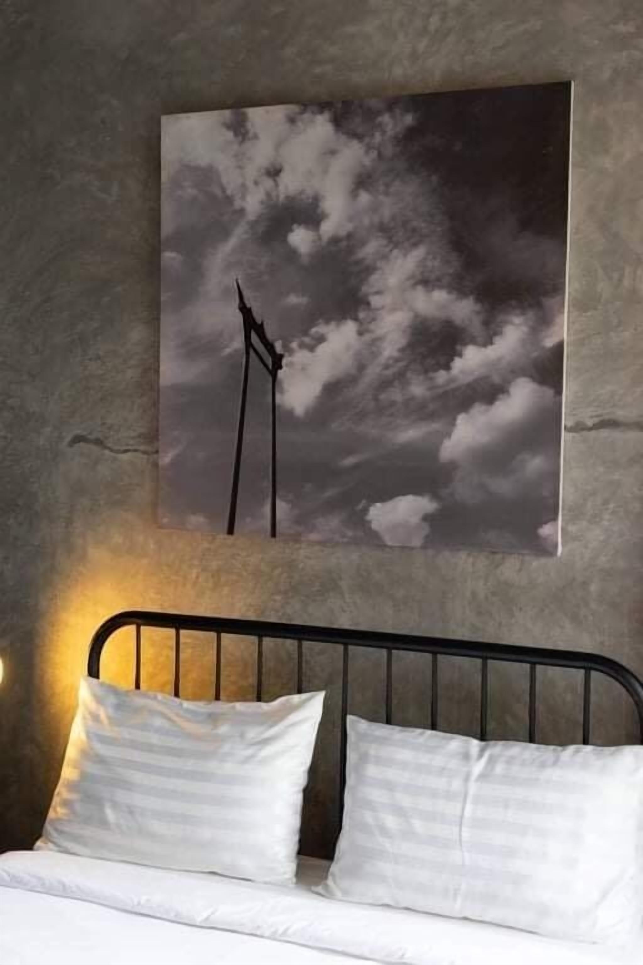 Una cama con ropa de cama blanca y cabecera de metal, un cuadro colgado en la pared que muestra un cielo nublado, y una lámpara de pared.