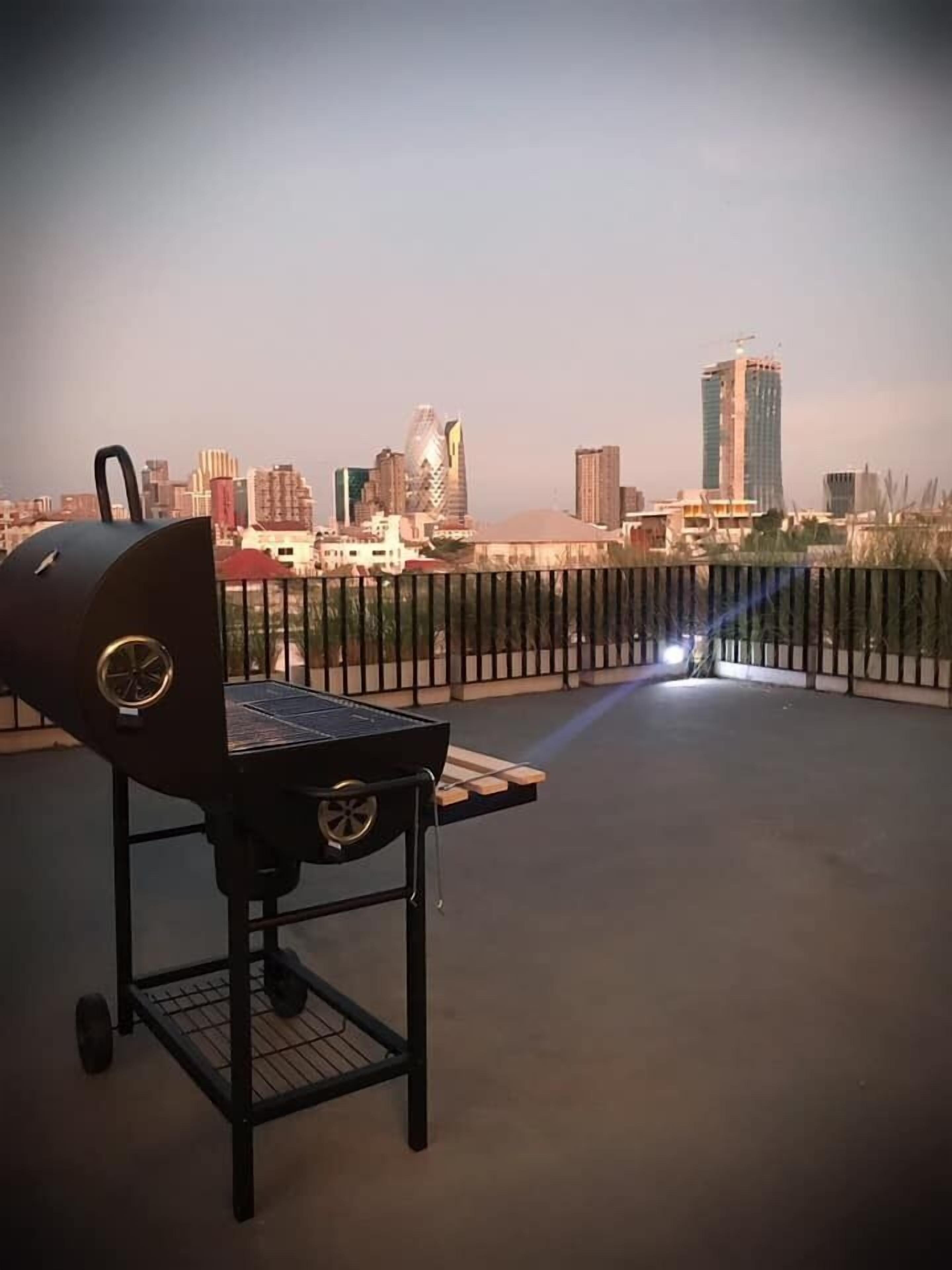 A rooftop with a barbecue grill, a metal railing, and a city skyline in the background.