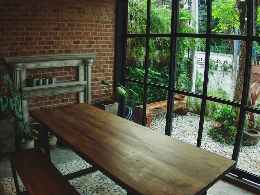 A rustic dining area with a wooden table and bench, brick walls, and large windows overlooking a garden.