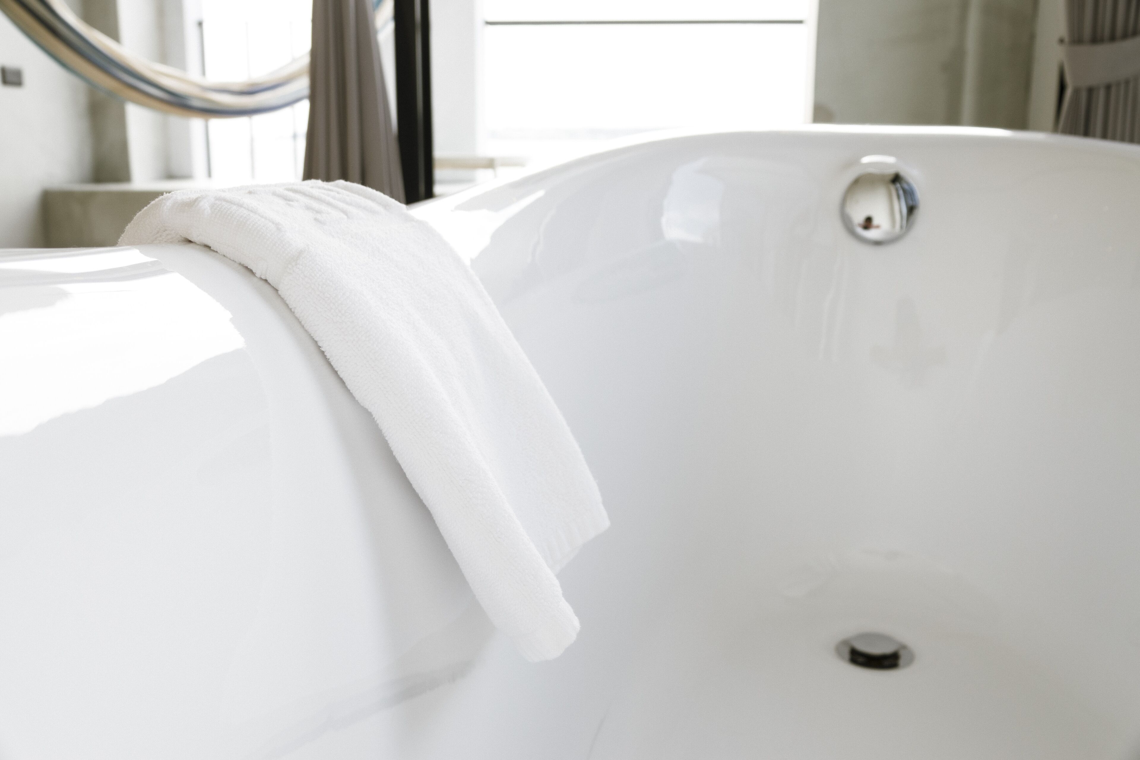Eine weiße Badewanne mit darübergelegtem Handtuch, einem sichtbaren Abfluss und einem Fenster im Hintergrund.