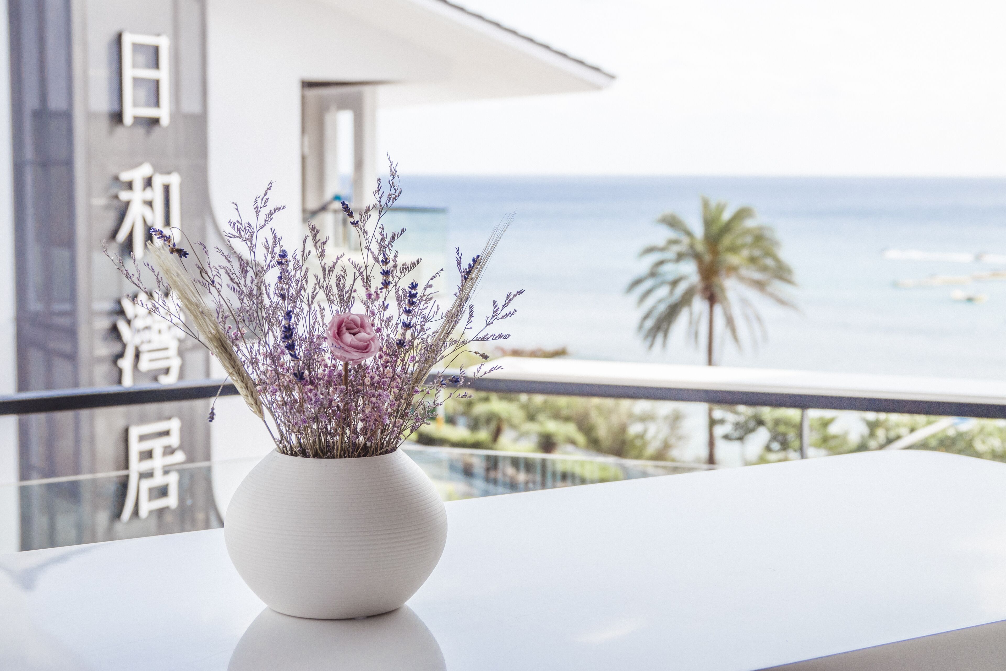 A white vase with purple flowers on a balcony overlooking the sea.