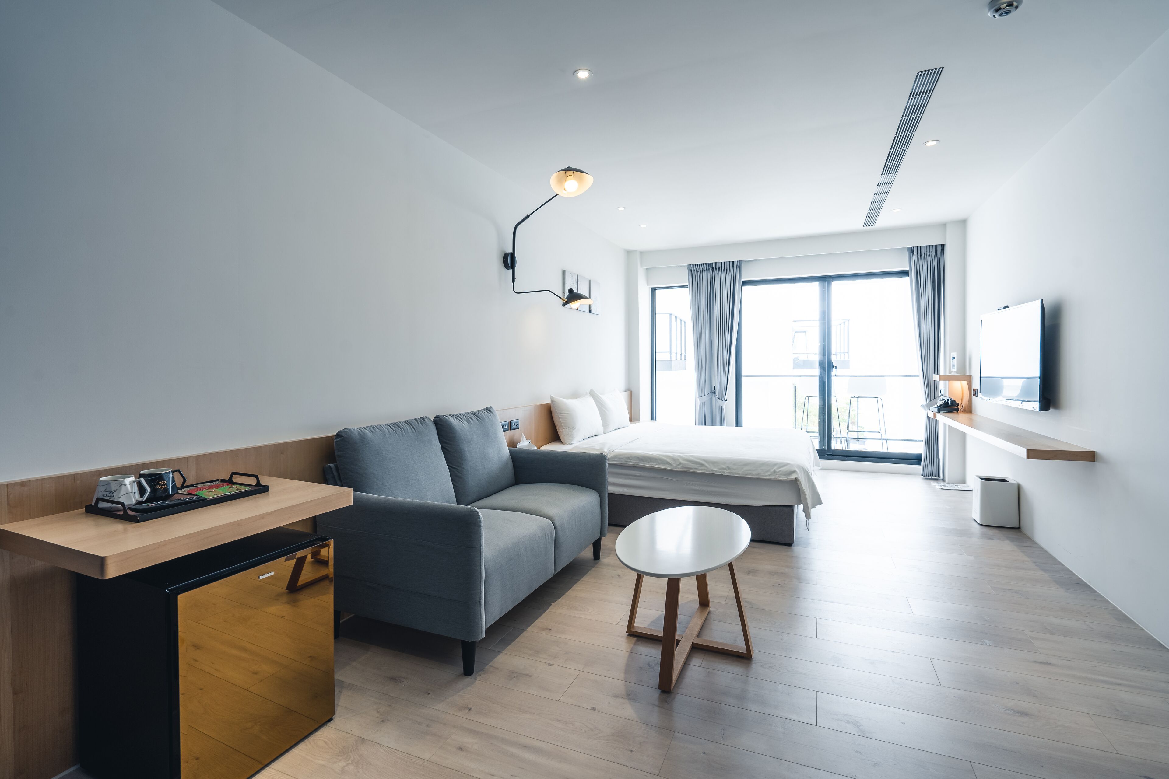 Una habitación de hotel moderna con sofá, una mesita, cama, televisor y ventana con cortinas.