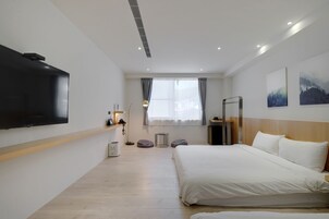 A modern hotel room with a large bed, a flat-screen TV mounted on the wall, a bedside table with a lamp, and a window with curtains.