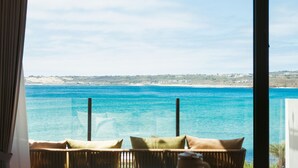 Vue depuis une pièce où se trouvent un fauteuil en osier et des coussins, donnant sur un paysage côtier avec une eau d’un bleu limpide et des collines lointaines.
