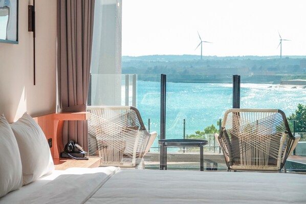 Habitació d'hotel amb un llit, dues cadires de vimetla, una taula petita i vistes al mar i aerogeneradors.