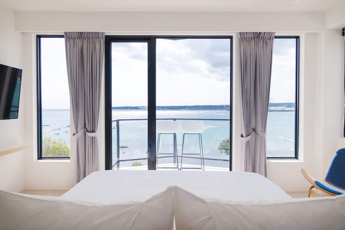 Un dormitorio con un ventanal que ofrece vistas al mar y a los barcos. Hay una cama, una silla y una mesa junto a la ventana.